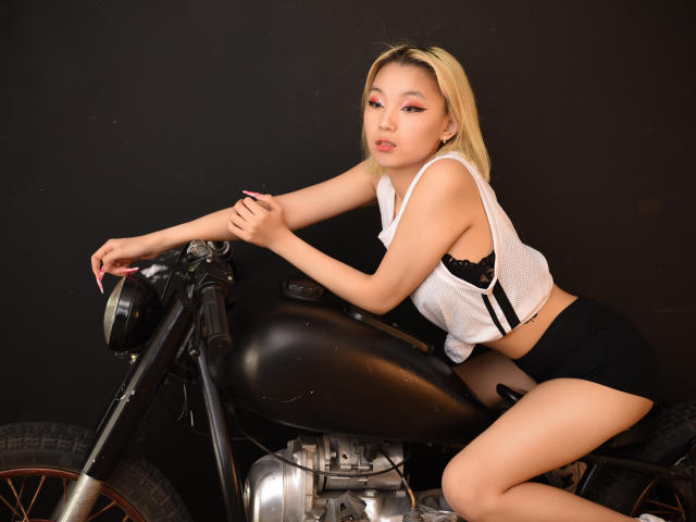 MillaWaves with blonde hair, white tank top, black shorts, and black bra, leaning on a black motorcycle, showing her ass, with pink eye makeup and a seductive look.