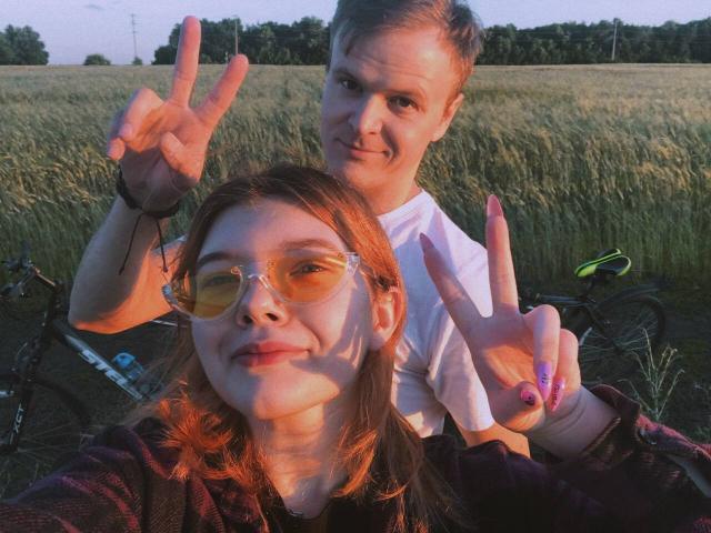 ChillOutt with glasses and orange-tinted lenses, making a peace sign, in front of ChillOutt with short hair, making a peace sign, in a field at sunset.