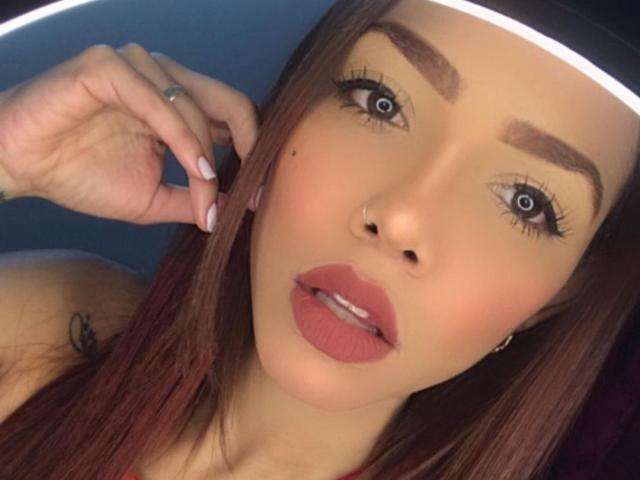 KaryHlovez a les cheveux bruns, les yeux bleus, des sourcils épais, du rouge à lèvres rose et une petite tache de rousseur sur sa joue droite. Sa main est près de son visage et elle porte une bague à son doigt..