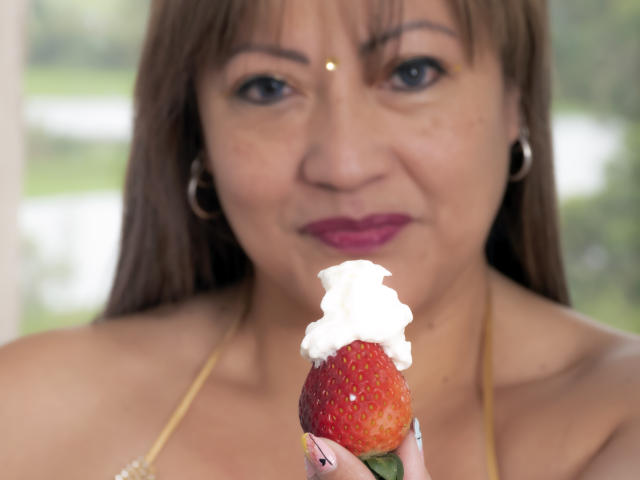 ChristinMoore with brown hair, pink lipstick, and hoop earrings, holding a strawberry with whipped cream in front of her smiling face, showing her tits and nose piercing.