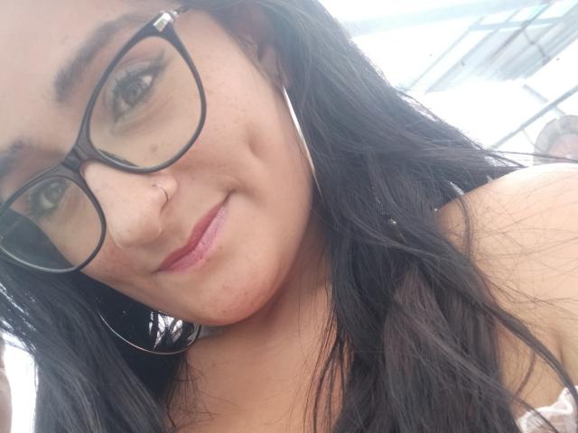 Meghan69 mit schwarzen Brillen, Nasenring und langen schwarzen Haaren, die leicht lächelt. Sie hat kleine Brüste, eine niedliche Nase und trägt große Reifenohrringe..