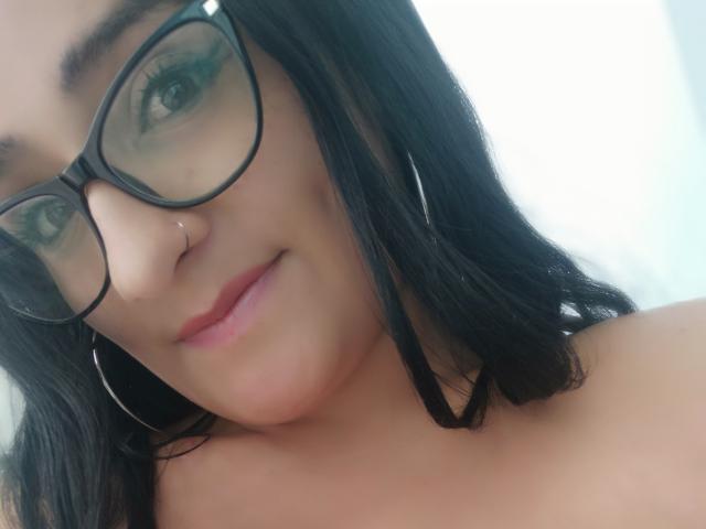 Meghan69 mit schwarzen Brillen, Nasenring und Ohrringen, lächelt in die Kamera, mit rosafarbenen Lippen und dunklem Haar. Brust nicht sichtbar, Fokus auf das Gesicht. Weiche, helle Haut..
