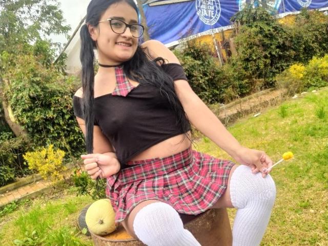 Meghan69 con lunghi capelli neri, occhiali, top corto nero, cravatta a quadretti rosa, gonna a quadretti rosa, calze bianche alte fino alla coscia, seduta su una panchina, che tiene in mano un lecca-lecca giallo e sorride..