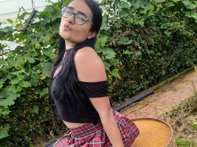 Meghan69 met lang zwart haar, een bril, een zwart top met een open schouder en een rode geruite rok, poseert met een gebogen rug en een speelse grijns, waarbij haar kleine borsten zichtbaar zijn..