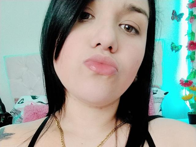 VivianaThomson with black hair, nose ring, pink lips, gold necklace, and tattoos, looking into camera with a pouty expression.