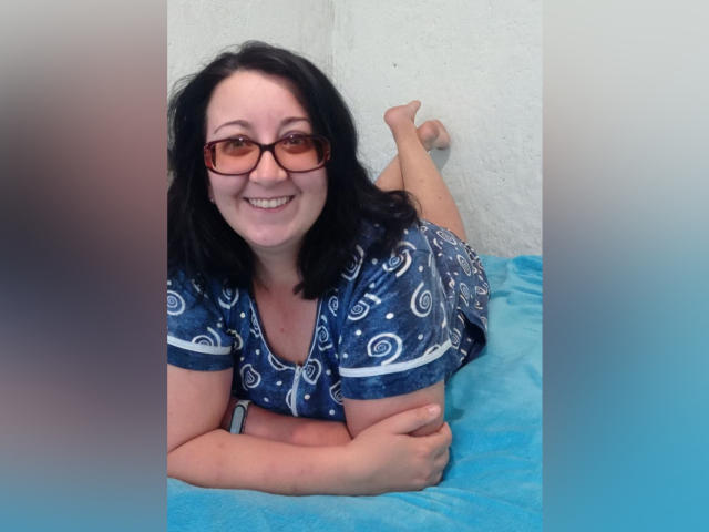 AnnaGoldXx lies on a blue blanket, smiling with glasses, black hair, and a blue patterned shirt, showing her bare ass and feet up against a white wall.