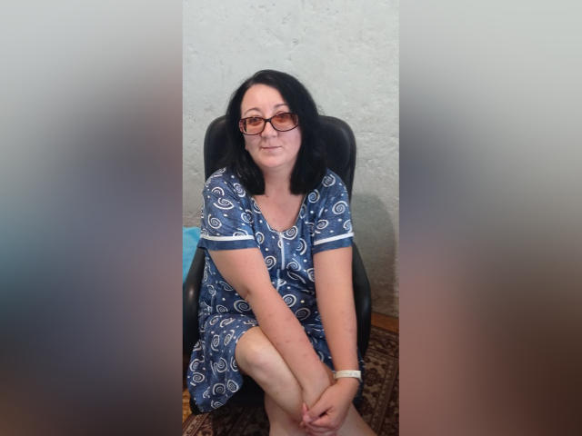 AnnaGoldXx is a chubby woman with black hair, glasses, and a blue patterned dress, sitting with her legs crossed, smiling slightly, wearing a white wristband.