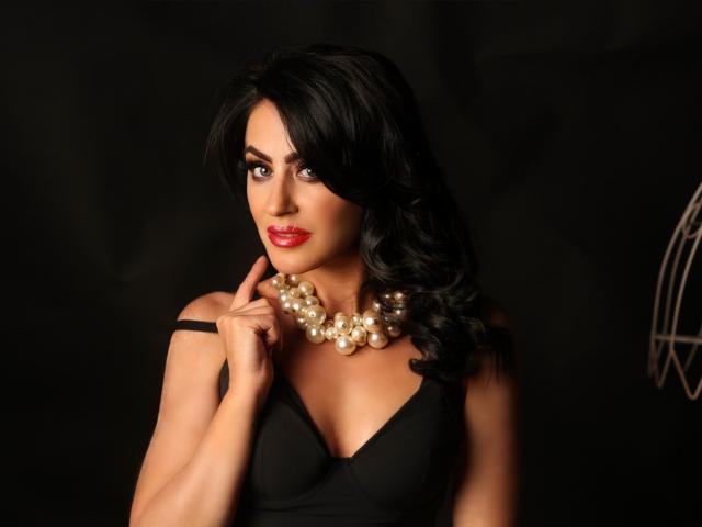 MistressTaylor with dark wavy hair, red lipstick, black dress, and pearl necklace, touching her chin, looking seductively at the camera, showing her tits in a dark setting.