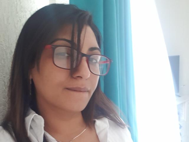 Jessyca, met een bril, bruin haar en een wit shirt, toont een lichte glimlach, heeft een schattig gezicht en een zilveren ketting, en staat bij een helder raam..