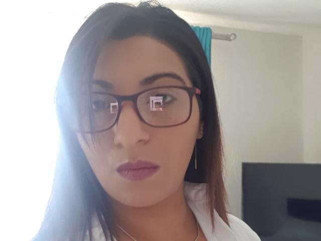 Jessyca a les cheveux bruns, des lunettes noires, du rouge à lèvres rose et une chemise blanche, avec un léger reflet lumineux sur le côté gauche de son visage. Ses fesses ne sont pas visibles..