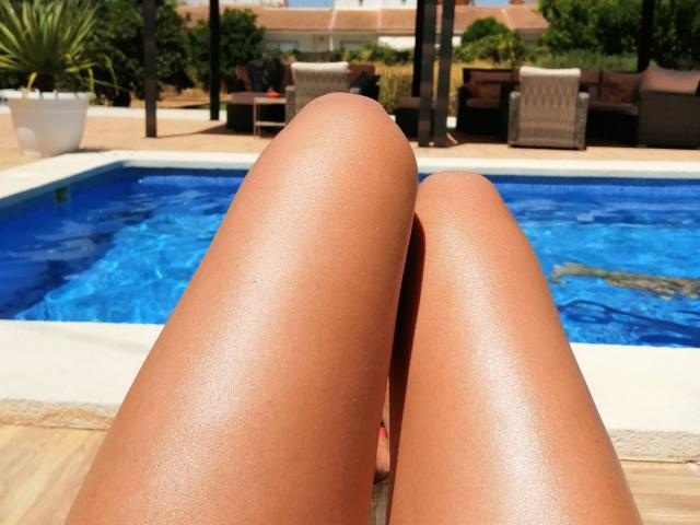 Les jambes lisses et bronzées de LaFrancaise, avec ses pieds dans une piscine, vues de son point de vue, montrant ses cuisses brillantes et bronzées, et de l'eau bleue en arrière-plan..