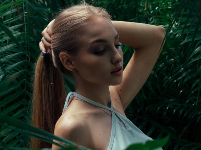 LinaCherry mit der Hand in ihrem langen, blonden Pferdeschwanz, die Augen geschlossen, trägt ein weißes Top ohne Ärmel und steht inmitten von grünem Laub, wobei ihre schlanken Arme und ihr dezentes Make-up zu sehen sind..