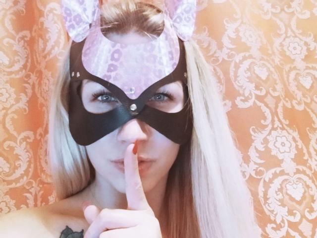 EqaJoe, con cabello rubio, máscara negra y un tocado de estampado de leopardo rosa, silenciando con el dedo en los labios, labios rosas, ojos azules y un tatuaje en su hombro derecho..