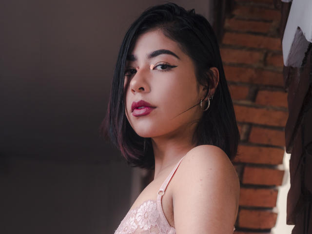 BiancaSpears heeft kort zwart haar, roze lippenstift en draagt een roze kanten bh. Ze kijkt schuin, met een vleugje eyeliner en gouden oorringen. Haar borsten zijn zichtbaar..