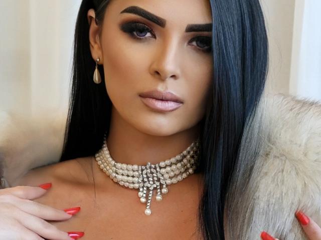 Devasha with long black hair, serious expression, wearing pearl necklace with dangling beads, fur collar, and red nails, looking directly at camera, tan skin, dark eyebrows.