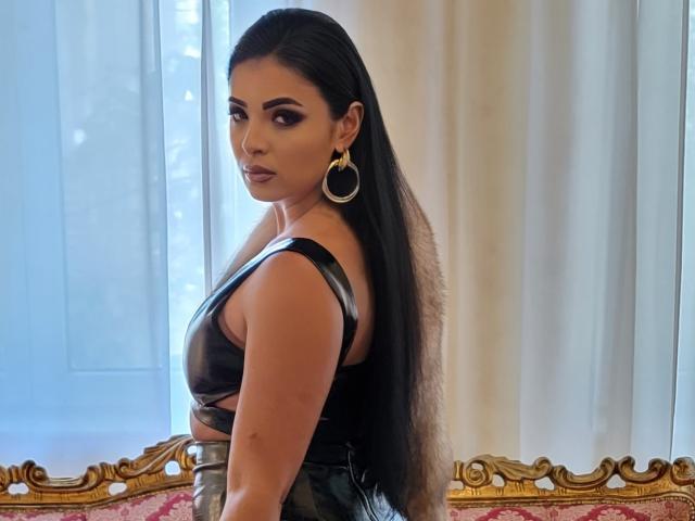 Devasha heeft lang zwart haar, draagt een glanzend zwart topje dat haar borsten laat zien, met gouden oorringen, en staat voor een raam, waarbij ze om zich heen kijkt..