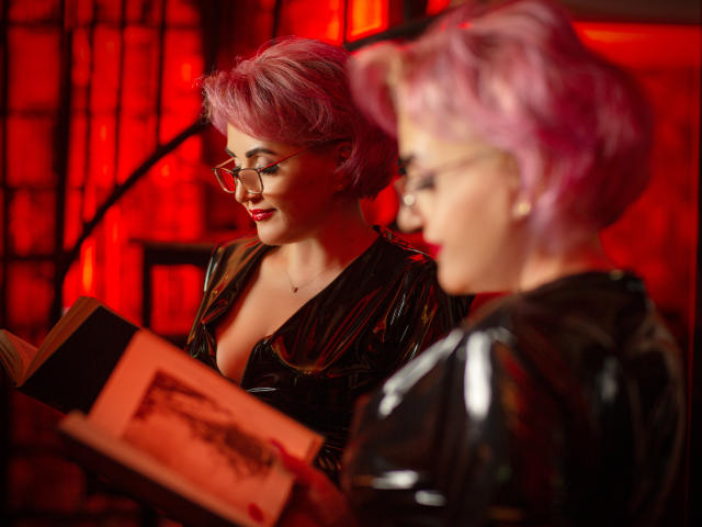 IsabelaConte with pink hair and glasses, in black latex, reading a book, showing tits, next to another IsabelaConte with pink hair, also in black latex, blurred in foreground.