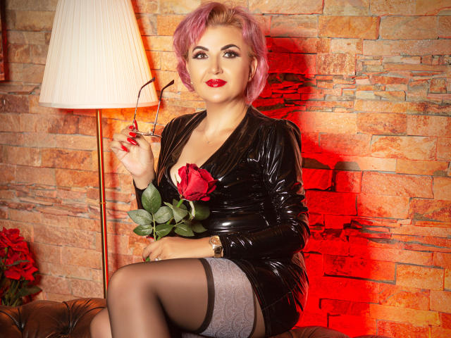 IsabelaConte with pink hair, red lipstick, black shiny dress, holding rose, glasses in hand, black stockings, sitting, brick wall, red light, leather chair, white lamp.