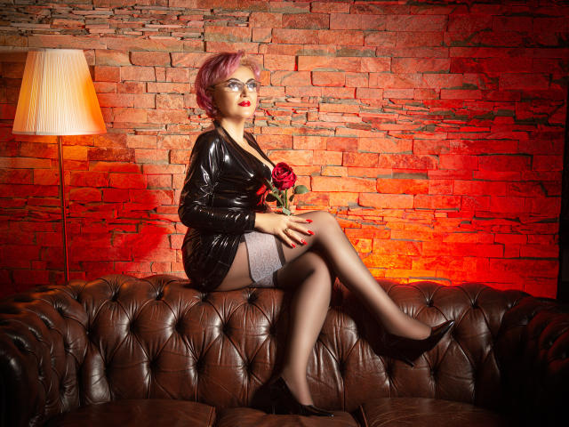 IsabelaConte sits on a brown leather couch, wearing a black latex dress, gray stockings, and black heels, with a red rose in her hand, her pink hair and glasses.