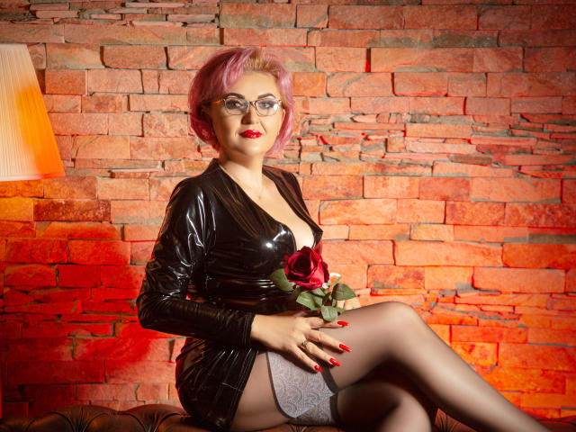 IsabelaConte with pink hair, glasses, red lipstick, black latex dress, holding red rose, crossed legs, gray lace panties, red nails, sitting on brown couch, brick wall background.