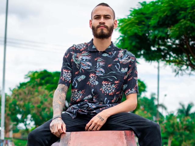 AndersX está sentado sobre un bloque rojo, vistiendo una camisa negra con estampado floral y pantalones negros, con un brazo tatuado y barba, y tiene una expresión seria. Su mano descansa sobre su rodilla..