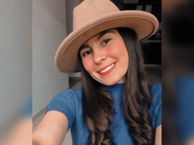 Alison Meyer, avec ses longs cheveux bruns, souriante, portant un chapeau beige et un t-shirt bleu côtelé, montre ses seins sur la photo. Son visage est mignon et elle a l'air heureuse..