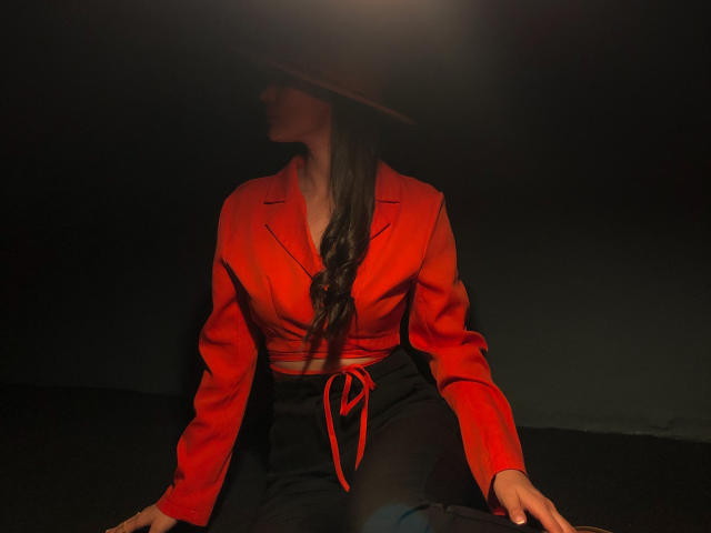Alison Meyer, vestindo um blazer vermelho, calças pretas e um chapéu marrom, sentada com cabelos castanhos longos, uma gravata vermelha e as mãos nos joelhos, sob uma iluminação fraca..