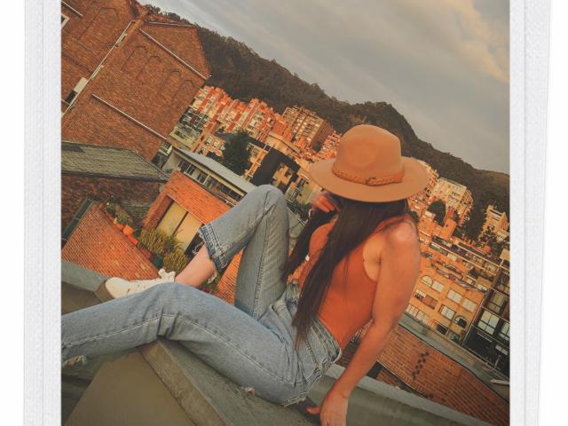 AlisonMeyer sits on a rooftop, topless, wearing a tan hat, blue jeans, and white socks. She has long brown hair, small tits, and is looking to the side.