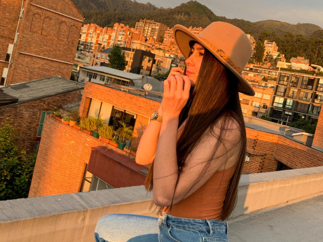 Alison Meyer, met lang bruin haar, draagt een beige hoed en een bruin top, praat aan de telefoon, zit op een dak, in het zonlicht, met een berg op de achtergrond, draagt jeans, houdt een telefoon vast, heeft een rustige uitdrukking..