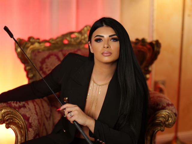 Devasha, con cabello negro largo, vestida con un blazer negro, cadenas doradas, y sosteniendo una batuta, sentada en una silla ornamentada de color rojo y dorado, luciendo segura de sí misma y atractiva..