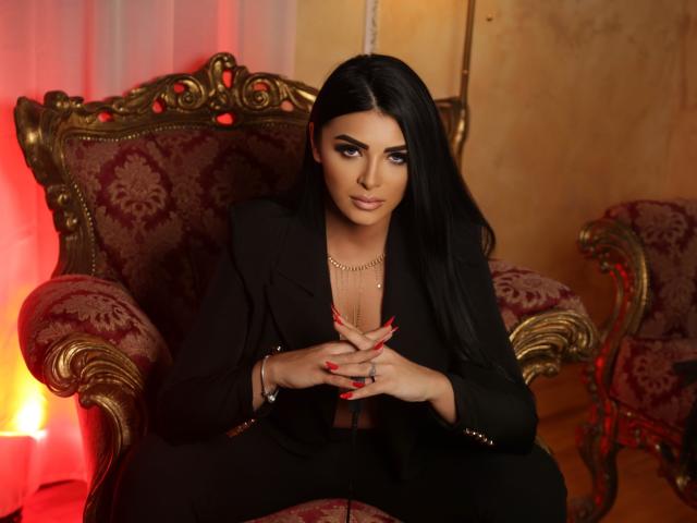 Devasha with long black hair, wearing a black suit, sitting on an ornate red and gold chair, hands with red nails, gold necklace, and looking at the camera.