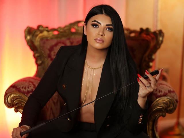 Devasha with long black hair, wearing a black blazer, gold necklace, and holding a whip, sitting on an ornate chair, her red nails contrasting with her tan skin.