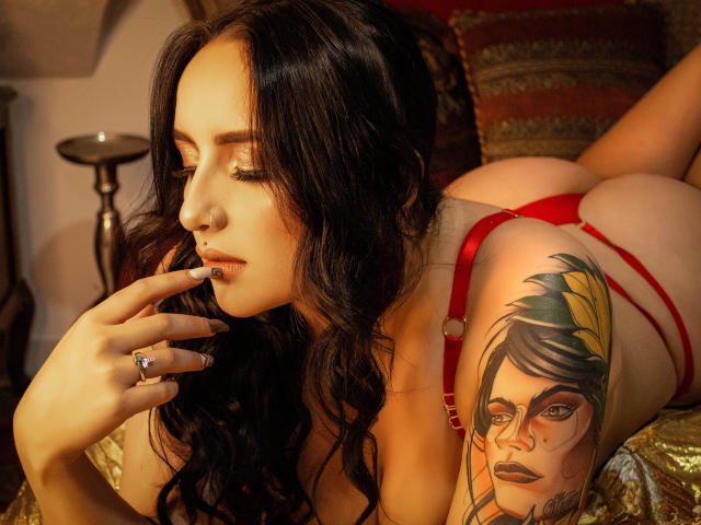 VioletSwitch, con lunghi capelli neri, biancheria intima rossa e un tatuaggio di una donna sul braccio, sdraiata su un letto, che si tocca le labbra, mostra il sedere e guarda in basso..