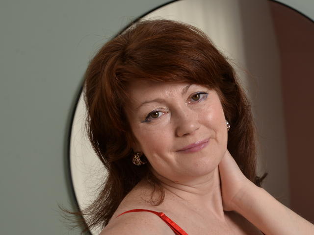 SadieLorri a les cheveux bruns, un léger sourire, et regarde l'appareil photo en posant sa main sur son cou. Elle porte un haut rouge et a de petites boucles d'oreilles..