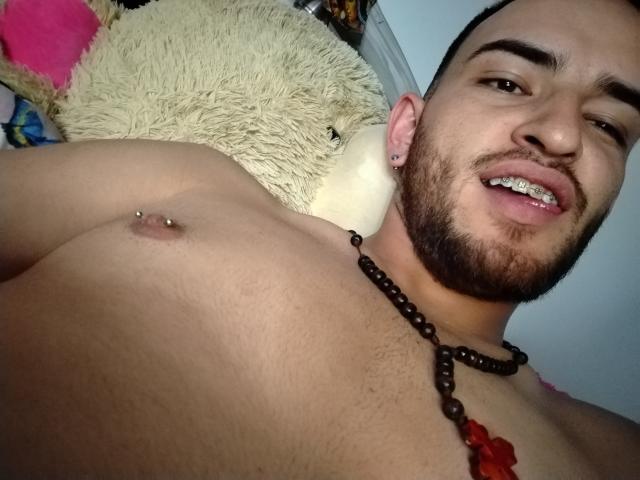 Jazmin e Ryan, senza camicia, con barba, capelli castani corti, sorridenti, che mostrano un capezzolo con piercing, collana di perline, fiore rosso, cuscino bianco, sopracciglia scure, occhi marroni, pelle chiara..