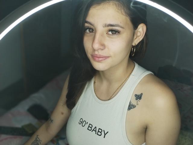 JazminYRyan hat eine hellbraune Haut, dunkelbraunes Haar, Ohrringe im Stecker-Stil, einen Nasenring und Tätowierungen an ihren Armen. Sie trägt ein weißes Tanktop mit der Aufschrift "90% BABY"..