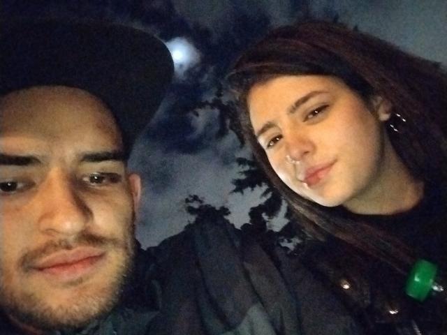 JazminYRyan has brown hair, a nose ring, and a subtle smile. JazminYRyan's boyfriend with a beard, hat, and slight smirk is on the left. Dark, moody background.
