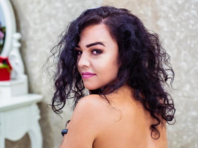 FunBrunettee with curly black hair, looking over her shoulder, showing her bare back and subtle makeup with pink lipstick, looking sexy and confident.