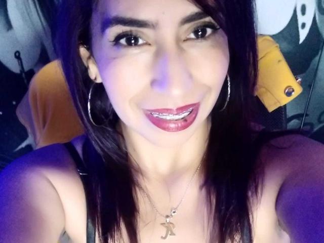 KatyKatt with dark brown hair, large hoop earrings, and shiny pink lipstick, smiling with tongue out, showing metal teeth, wearing a black strap top, and a small pendant.