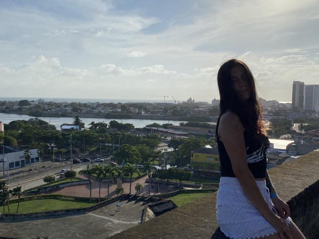 JuliaGarciax with long dark hair, black sleeveless top, and white lace skirt, standing on a rooftop, sunlight on her face, city and river in the background.