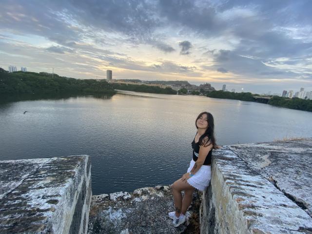 JuliaGarciax with long black hair, black top, white skirt, and white shoes, sitting on a weathered stone ledge, looking up, with a calm river and cloudy sky in background.