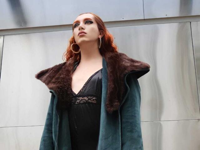Julia Fanning, con cabello rojo, vestida con un abrigo verde de terciopelo con detalles de piel marrón, una blusa negra con encaje y grandes aros, mirando hacia arriba con confianza..
