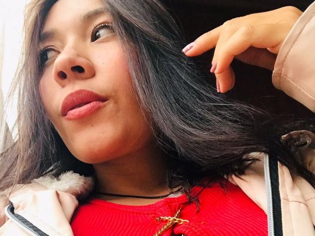 HelenRoss has dark, wavy hair, red lipstick, and a slight smile. She's wearing a pink jacket, red top, and a gold necklace with a star-shaped pendant.