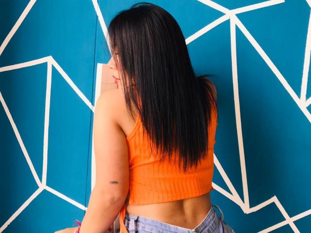 Helen Ross, com cabelos longos e pretos, blusa cropped laranja texturizada e jeans azuis, exibindo uma tatuagem no braço direito, apoiada em uma parede verde-água com linhas geométricas brancas..