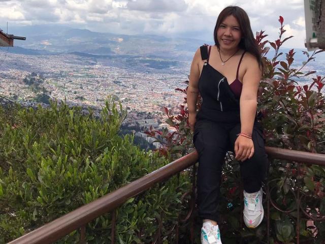 Helen Ross, con il seno scoperto, indossa una maglietta nera con una spalla scoperta, è seduta su una ringhiera, sorride, e indossa scarpe da ginnastica bianche con dettagli blu, circondata da vegetazione verde e rossa..