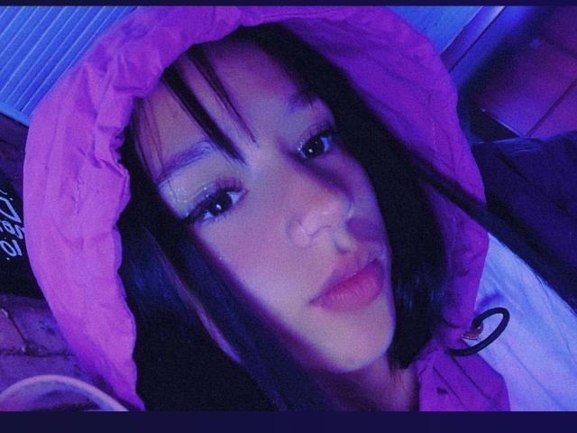 KloeThompson with dark hair, pink hooded jacket, purple eyeshadow, nose piercing, and blue-tinted face, looking at camera with slightly parted lips.