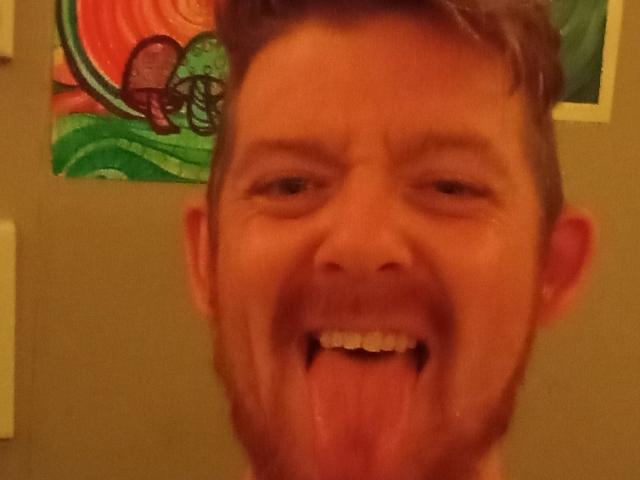 Le visage de FreddyMac est sur une photo où il a la langue tirée et fait une expression ridicule, presque stupide. Il a une barbe et des cheveux bruns courts, et il a l'air idiot..