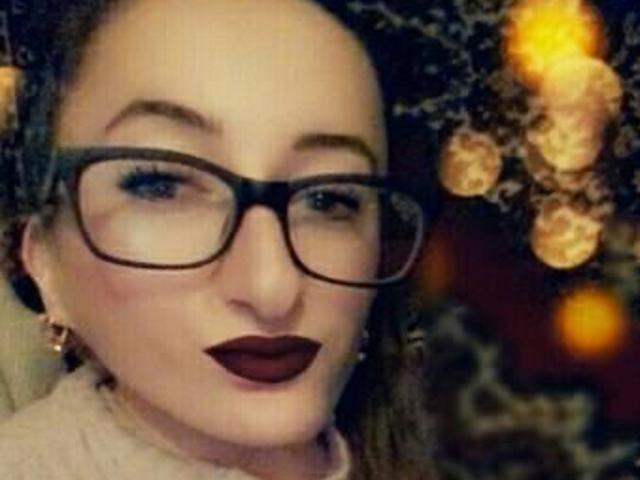 JollyHot69 con lápiz labial burdeos oscuro, gafas de montura negra y cabello castaño rizado, usando aretes dorados, mirando a la cámara con una sonrisa sutil, en un fondo suavemente iluminado y borroso..