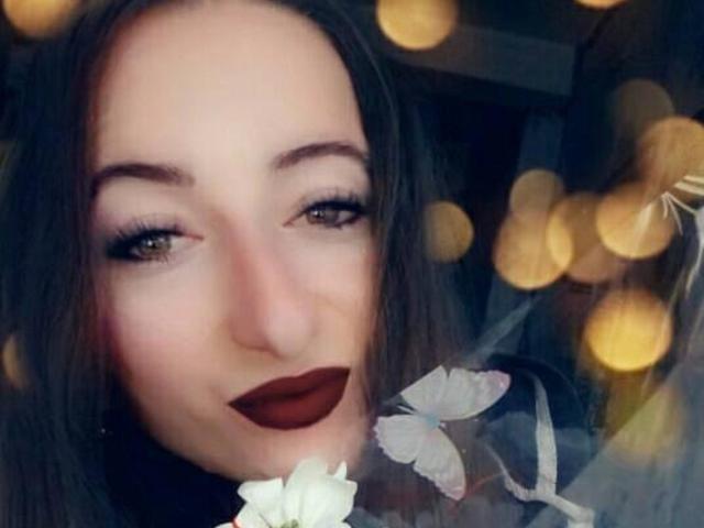 JollyHot69 mit dunklem Lippenstift, grünen Augen und schwarzen Augenbrauen, hat ein sanftes Lächeln, im Hintergrund sind Bokeh-Lichter zu sehen, und trägt ein Blumenmuster-Kleidungsstück..