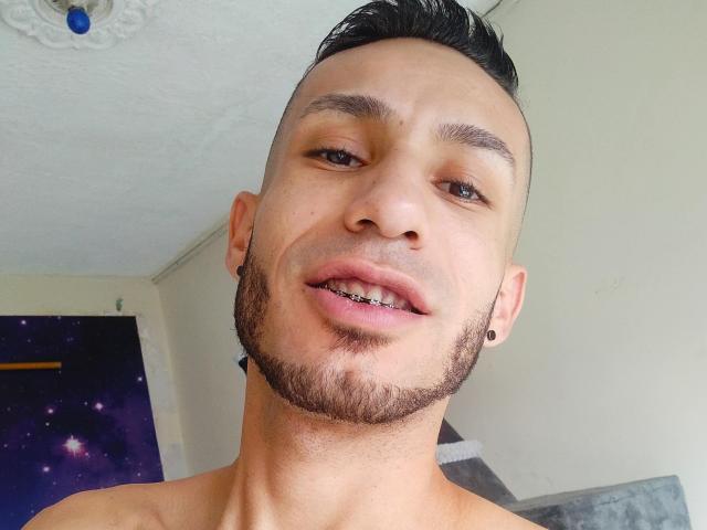 WolfMonsterCock ha una carnagione marrone chiaro, capelli neri corti, una barba, piercing alle orecchie neri e denti di metallo. È senza camicia, con la bocca leggermente aperta e un'espressione rilassata..